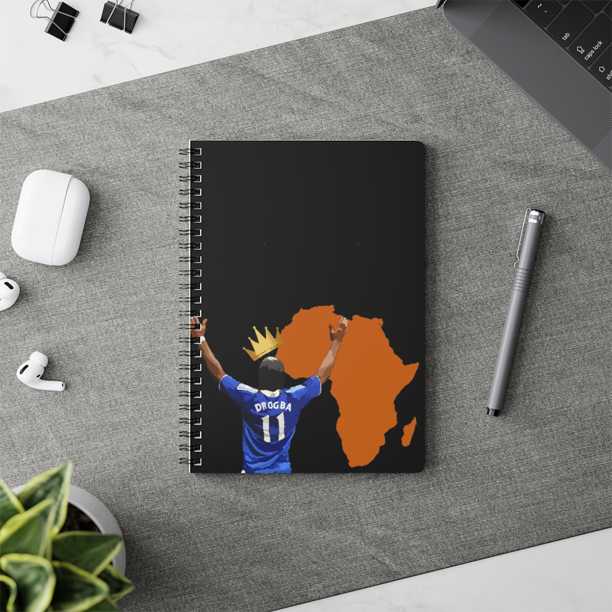 Contextual Front view of Chelsea Didier Drogba with a crown a top his head with the orange coloured continent of Africa on an A5 spiral notepad on top of a desk.